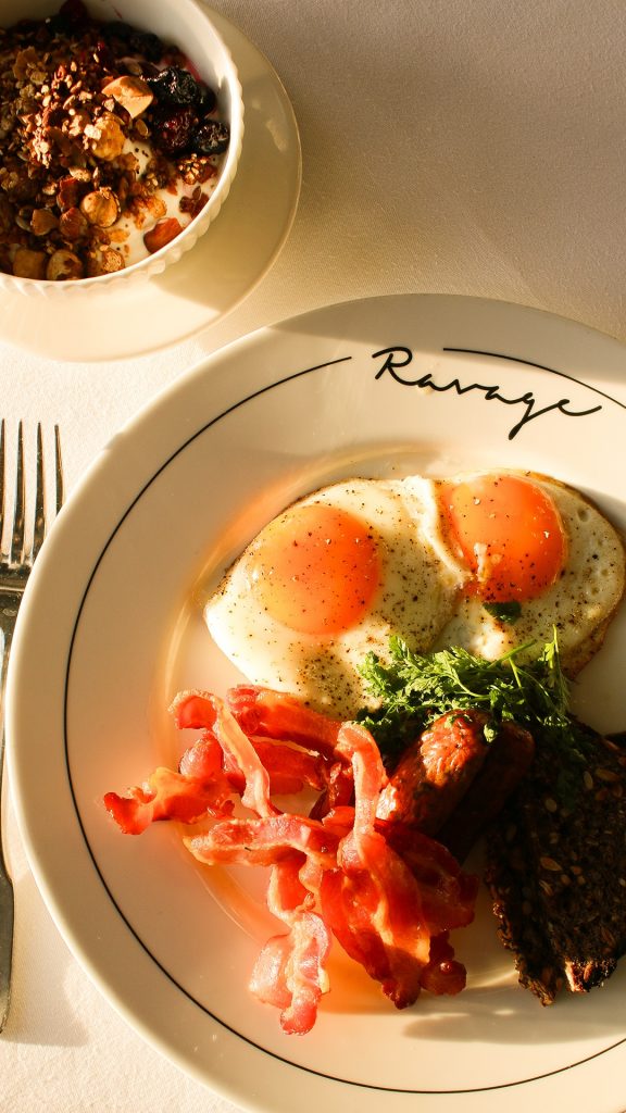 Morgenmad på Ravage med spejlæg, bacon, pølser, rugbrød og granola i varmt morgensollys.